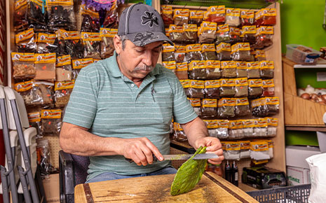 man cutting a prickly pear cactus in front of spices in his store